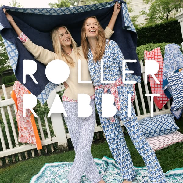 Two women are outside on a sunny day modeling Roller Rabbit pajamas. A blanket is held over their heads, and a display o