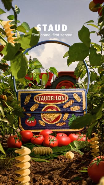 This image features a Staud purse designed to look like pasta packaging. The purse is presented outdoors amongst tomato 