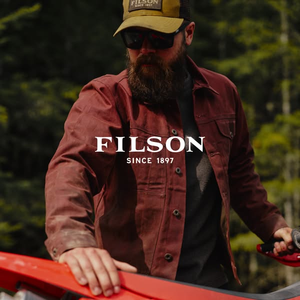 A close up shot of a bearded man wearing a Filson jacket and hat, with the Filson logo laid over the image. The image is
