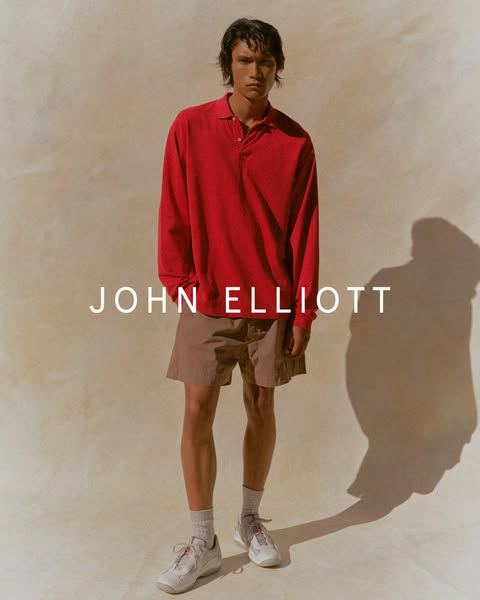 An image ad promoting John Elliott clothing. A male model is standing in front of a tan wall wearing a red long sleeve p