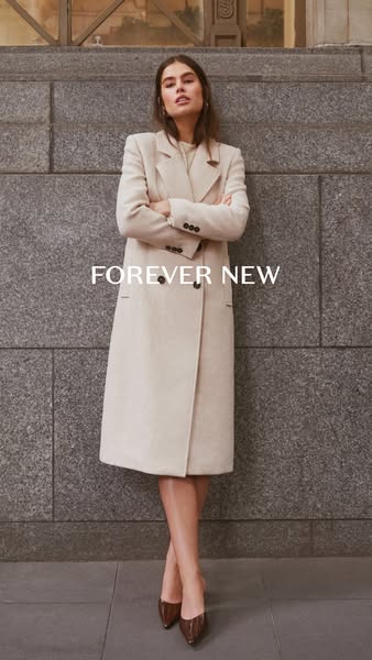 A woman models a Forever New coat in front of a stone wall.