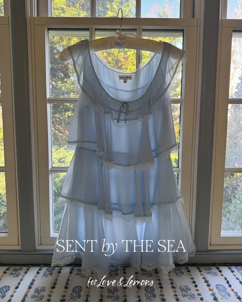 This is an image ad for Love & Lemons that shows a tiered baby blue dress with lace trim hanging in front of a windo